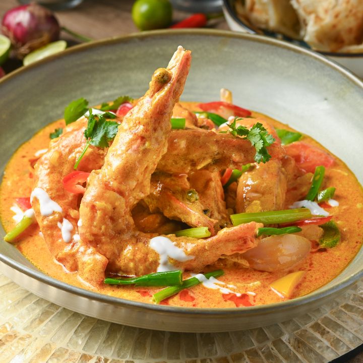Tiger prawns in light yellow curry sauce, served in ceramic bowl surrounded by red onion, chillies and limes.