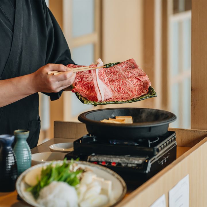 Chef in black placing wagyu beef slice on green dish into cast iron sukiyaki pot with chopsticks.